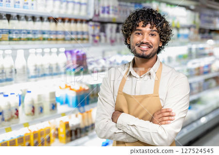 A cheerful store employee stands with folded arms, smiling in front of the dairy section filled with products. The grocery store worker looks confident and ready to assist. 127524545