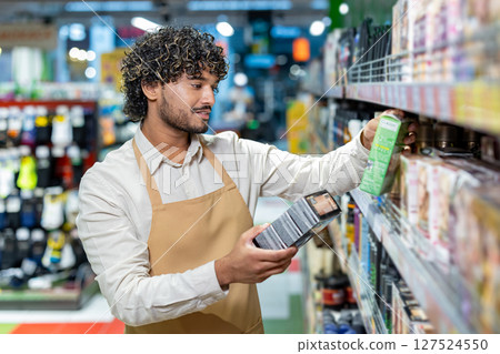 A friendly worker, wearing an apron, is reviewing products on the shelf at the store. He carefully considers the labels. A friendly worker, wearing an apron, is reviewing products on the shelf at the store. He carefully considers the labels. 127524550