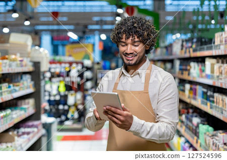 A smiling store clerk checks inventory with a tablet in a grocery store setting. Shelves filled with products surround the employee. A smiling store clerk checks inventory with a tablet in a grocery store setting. Shelves filled with products surround the employee. 127524596