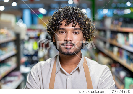 A portrait of a store worker standing between shelves with a serious expression. He wears a light shirt and an apron, looking directly at the camera. A portrait of a store worker standing between shelves with a serious expression. He wears a light shirt and an apron, looking directly at the camera. 127524598