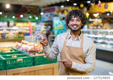 A smiling grocery store employee points to the fresh produce section, offering a friendly and inviting experience for customers in this environment. A smiling grocery store employee points to the fresh produce section, offering a friendly and inviting experience for customers in this environment. 127524605