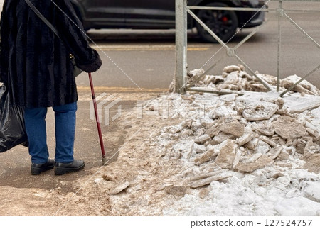 An unknown woman with a cane is standing waiting for the green light of a traffic light. An individual confidently navigating a snowy sidewalk, showcasing resilience and strength amid winters many 127524757