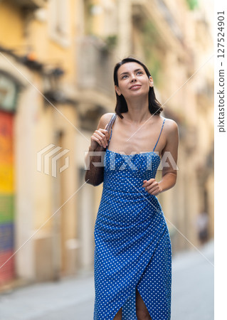 Cheerful brunette girl in blue dress walking along the street among architecture with bag Cheerful brunette girl in blue dress walking along the street among architecture with bag 127524901