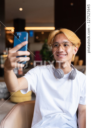 Asian teenager man with blonde hair inside cafe restaurant taking selfie with phone 127525645