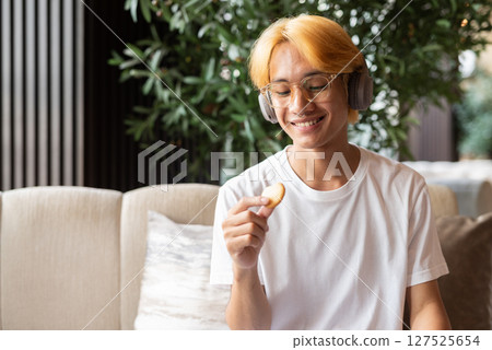 Asian teenager man with blonde hair inside cafe restaurant eating cookie Asian teenager man with blonde hair inside cafe restaurant eating cookie 127525654