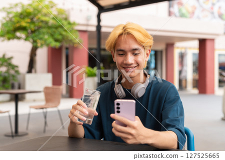 Asian gay teenager drinking water and using phone outdoors in cafe restaurant 127525665