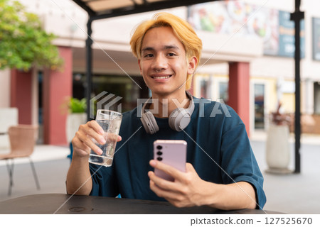 Asian gay teenager drinking water and using phone outdoors in cafe restaurant 127525670