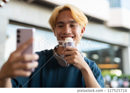 Young man eating soft serve ice cream outdoors while taking selfie with phone Young man eating soft serve ice cream outdoors while taking selfie with phone 127525718