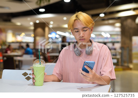 Thai gay teen man with headphones using phone and drinking iced latte green tea in modern cafe Thai gay teen man with headphones using phone and drinking iced latte green tea in modern cafe 127525735