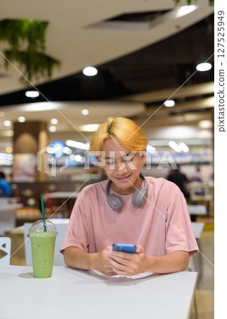 Thai gay teen man with headphones using phone and drinking iced latte green tea in modern cafe 127525949