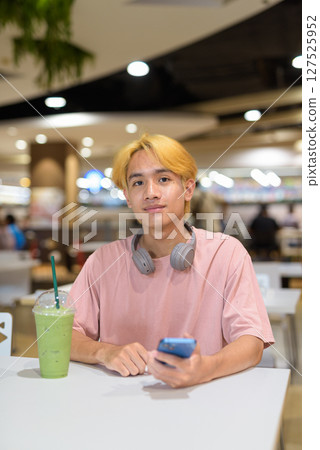 Thai gay teen man with headphones using phone and drinking iced latte green tea in modern cafe Thai gay teen man with headphones using phone and drinking iced latte green tea in modern cafe 127525952