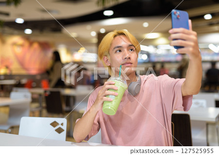 Thai gay teen man with headphones using phone and drinking iced latte green tea in modern cafe Thai gay teen man with headphones using phone and drinking iced latte green tea in modern cafe 127525955