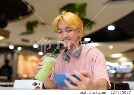 Thai gay teen man with headphones using phone and drinking iced latte green tea in modern cafe Thai gay teen man with headphones using phone and drinking iced latte green tea in modern cafe 127525957