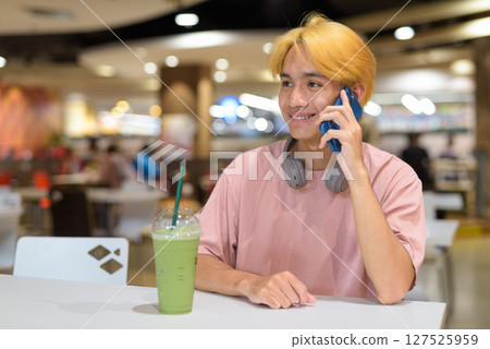 Thai gay teen man with headphones using phone and drinking iced latte green tea in modern cafe 127525959