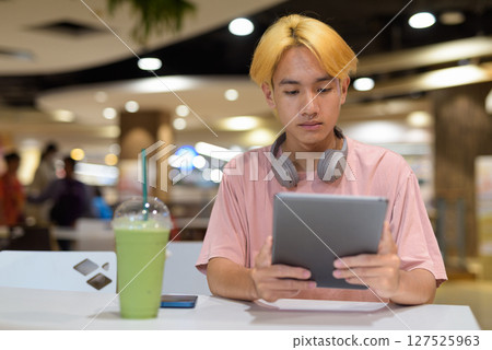 Thai gay teen man with headphones using digital tablet and drinking iced latte green tea Thai gay teen man with headphones using digital tablet and drinking iced latte green tea 127525963