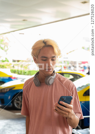 Young Asian gay teen tourist using phone outdoors with taxis behind Young Asian gay teen tourist using phone outdoors with taxis behind 127526020