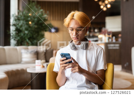 Asian teenager man with blonde hair inside cafe restaurant using phone Asian teenager man with blonde hair inside cafe restaurant using phone 127526021
