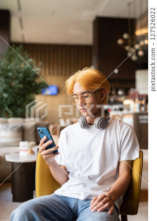 Asian teenager man with blonde hair inside cafe restaurant using phone 127526022