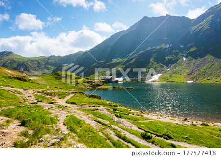 balea lake of romania. alpine landscape of fagaras mountains with pond. sunny weather with clouds on the blue sky above carpathians. snow and grass on the hillside in summer. calm water surface 127527418