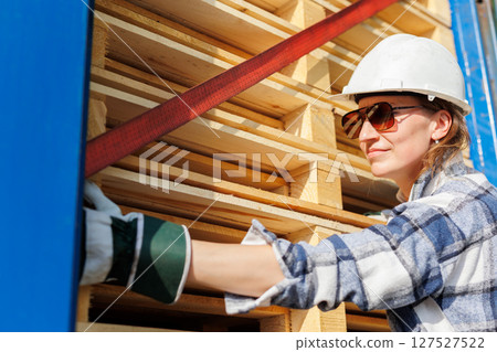 Portrait happy female driver wearing sunglasses and safety helmet smiles in front of blue truck and stacked pallets. Female person girl professional job career working driving freight engine lorry Portrait happy female driver wearing sunglasses and safety helmet smiles in front of blue truck and stacked pallets. Female person girl professional job career working driving freight engine lorry 127527522