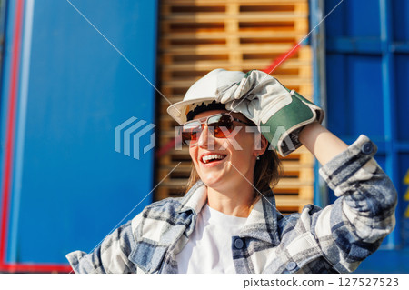 Portrait happy female driver wearing sunglasses and safety helmet smiles in front of blue truck and stacked pallets. Female person girl professional job career working driving freight engine lorry Portrait happy female driver wearing sunglasses and safety helmet smiles in front of blue truck and stacked pallets. Female person girl professional job career working driving freight engine lorry 127527523
