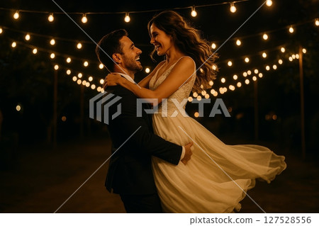 Groom lifting bride in wedding dress under string lights at night during outdoor evening celebration Groom lifting bride in wedding dress under string lights at night during outdoor evening celebration 127528556