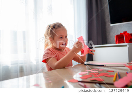 Portrait of cute little Caucasian girl smiling is sitting at a table and cutting out paper hearts. Child learns how to make crafts for Mother's Day and Valentine's Day 127529449