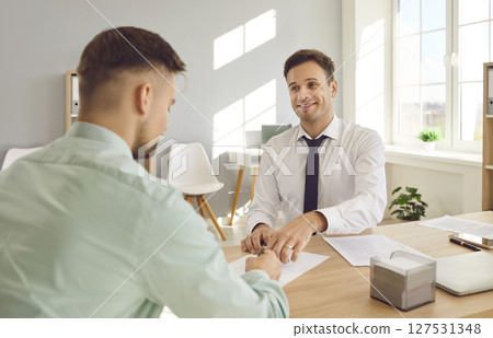 Two smiling business men at work in office talking, reaching agreement and signing contract. 127531348