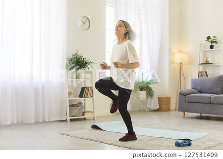 Smiling mature woman running in place in living room, following video tutorial for home workout. 127531409