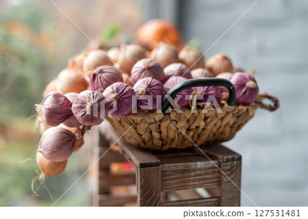 A wicker basket brimming with fresh red and brown onions, resting on a wooden stool in a cozy and natural setting, showcasing organic produce and kitchen essentials. A wicker basket brimming with fresh red and brown onions, resting on a wooden stool in a cozy and natural setting, showcasing organic produce and kitchen essentials. 127531481