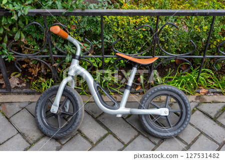 A compact metal balance bike for children parked against a wrought iron fence in a neatly landscaped area with paving and greenery. 127531482