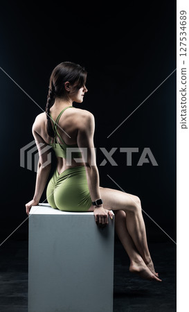 Athletic woman sitting on white cube in sportswear posing on black background Athletic woman sitting on white cube in sportswear posing on black background 127534689