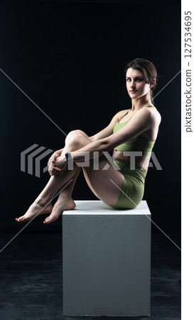 Athletic woman sitting on white cube in sportswear posing on black background Athletic woman sitting on white cube in sportswear posing on black background 127534695