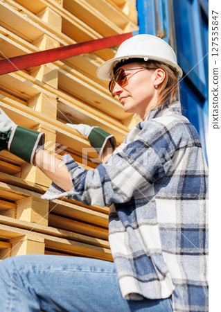 Portrait happy female driver wearing sunglasses and safety helmet smiles in front of blue truck and stacked pallets. Female person girl professional job career working driving freight engine lorry Portrait happy female driver wearing sunglasses and safety helmet smiles in front of blue truck and stacked pallets. Female person girl professional job career working driving freight engine lorry 127535847