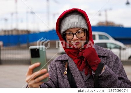 Smiling woman in winter attire takes a selfie outdoors on a chilly day near urban construction site 127536074