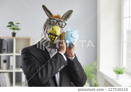 Funny man in a suit wearing animal donkey mask on head standing in the office holding piggy bank. 127536510