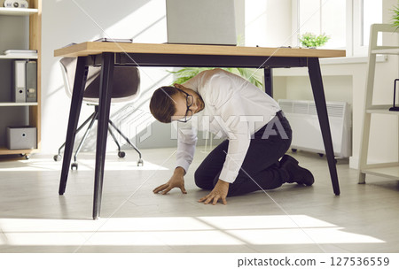 Scared young office worker hiding under his working desk during dangerous earthquake Scared young office worker hiding under his working desk during dangerous earthquake 127536559