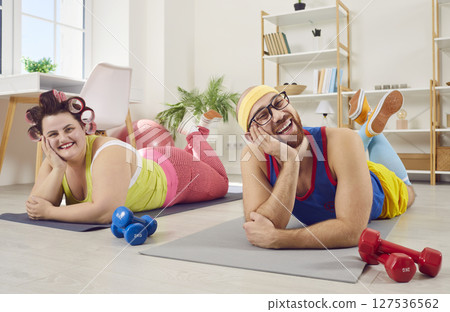 Funny couple in bright sportswear doing sport exercises at home lying on yoga mat with dumbbells. 127536562