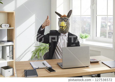 Funny business man wearing animal mask sitting at the desk showing thumb up sign on his workplace. Funny business man wearing animal mask sitting at the desk showing thumb up sign on his workplace. 127536572