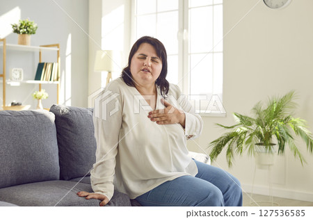 Overweight woman with heart disease sitting on sofa and suffering from pain in her chest 127536585