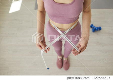 Top view of sporty woman in sportswear measure waist with measuring tape at home. 127536593