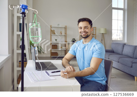 Happy young man receiving intravenous vitamin infusion while sitting at working desk at home 127536669