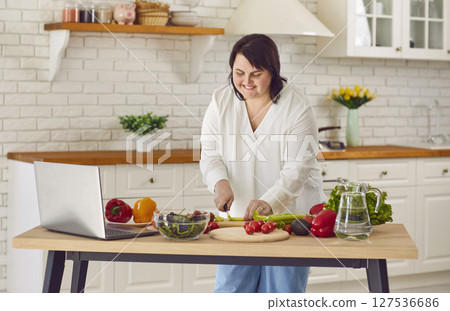 Portrait of fat overweight young woman preparing fresh vegetable salad looking at recipes on laptop 127536686