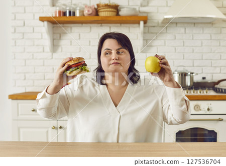 Overweight woman holding apple and burger and choosing between healthy and unhealthy food Overweight woman holding apple and burger and choosing between healthy and unhealthy food 127536704
