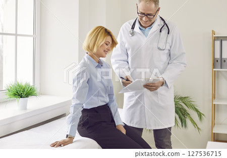 Mature male doctor talking with young woman who sitting on couch in medical office Mature male doctor talking with young woman who sitting on couch in medical office 127536715