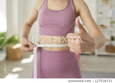 Young woman who has lost some weight measures her thin waist and gives a thumbs up 127536722