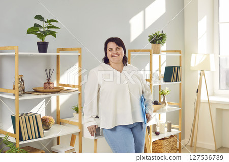 Portrait of a fat plus size brunette woman standing at home holding folder in hands and smiling. Portrait of a fat plus size brunette woman standing at home holding folder in hands and smiling. 127536789