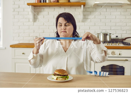 Fat overweight young woman sitting on kitchen holding a measuring tape with a burger on the table. Fat overweight young woman sitting on kitchen holding a measuring tape with a burger on the table. 127536823