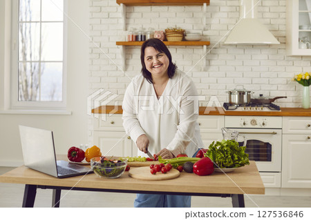 Fat overweight woman preparing fresh vegetable salad looking at recipes on laptop and smiling. 127536846