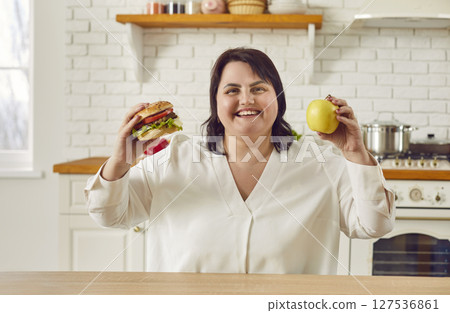 Young overweight plus size fat woman holding a burger in one hand and an apple in other. Young overweight plus size fat woman holding a burger in one hand and an apple in other. 127536861